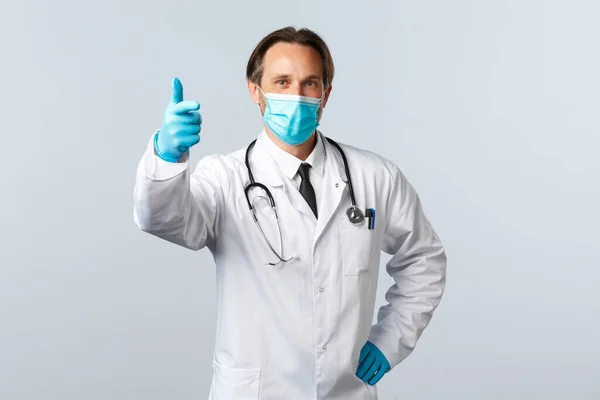 Covid-19, virüsü önleme, sağlık çalışanları ve aşı konsepti. Tıbbi maske ve eldiven giymiş tatmin olmuş bir doktor, baş parmağını kaldır, iyi bir jest yap, reklam reklamından memnun ol. — Stok fotoğraf