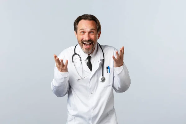 Covid-19, ξέσπασμα του ιού της στέψης, επαγγελματίες υγείας και πανδημία. Ενθουσιασμένος ευτυχισμένος αρσενικό γιατρό γελώντας και εξηγώντας κάτι αστείο, χαμογελώντας ευρέως, στέκεται λευκό φόντο — Φωτογραφία Αρχείου