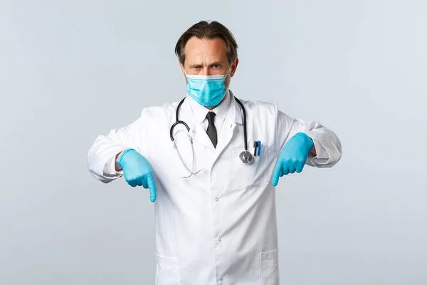 Covid-19, πρόληψη του ιού, εργαζόμενοι στον τομέα της υγείας και την έννοια του εμβολιασμού. Σοβαρός αποφασισμένος γιατρός που δείχνει μέτρα κατά του κορωναϊού, φοράει ιατρική μάσκα και γάντια, δείχνοντας τα δάχτυλα προς τα κάτω — Φωτογραφία Αρχείου