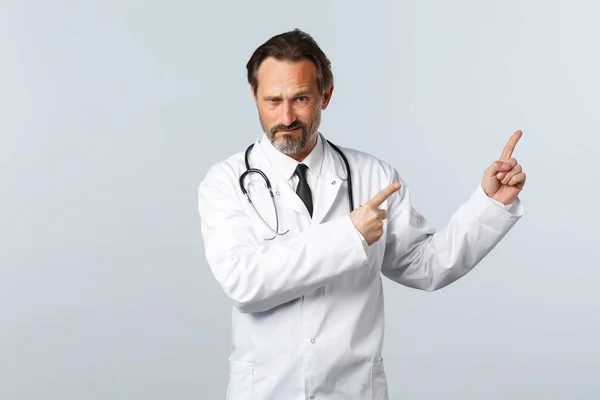 Covid-19, coronavirus outbreak, healthcare workers and pandemic concept. Skeptical and unsure male doctor, physician in white coat, frowning doubtful and pointing upper right corner — Stock Photo, Image