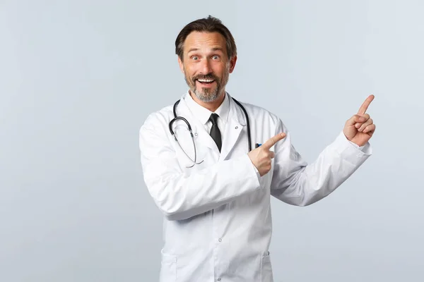 Covid-19, epidemia koronawirusów, pracownicy służby zdrowia i koncepcja pandemii. Szczęśliwy entuzjastyczny lekarz w białym płaszczu, skierowany w prawym górnym rogu na baner, uśmiechnięty, zapraszający pacjentów do kliniki — Zdjęcie stockowe