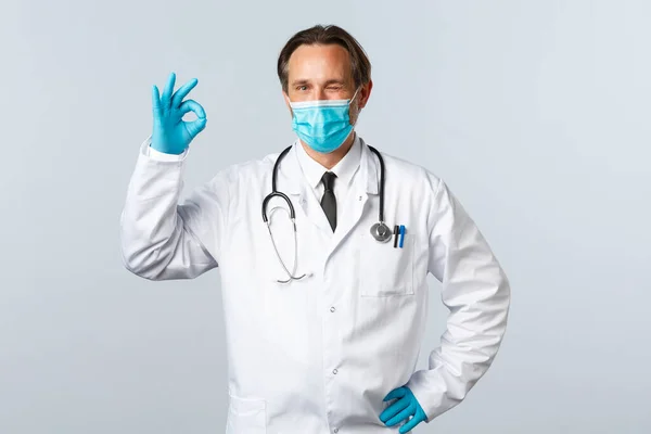 Covid-19: 바이러스 예방, 의료 종사자 및 예방 개념. 의료용 마스크와 장갑을 끼고 윙크를 하고 좋은 표시를 보이며 즐거움에 넘쳐 있는 의사는 품질 과 흰색 배경을 보장 한다 — 스톡 사진