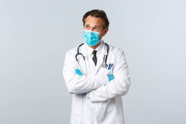 Covid-19, virüsü önleme, sağlık çalışanları ve aşı konsepti. Tıbbi maskeli ve eldivenli sorunlu doktor, sol üst köşeye bakıyor endişeli, kararını düşünüyor.