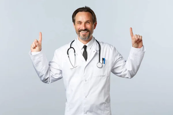 Covid-19, coronavirus outbreak, healthcare workers and pandemic concept. Cheerful smiling male doctor in white coat, pointing fingers up to show banner, promote product medication — Stock Photo, Image