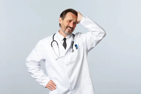 Covid-19, ξέσπασμα του ιού της στέψης, επαγγελματίες υγείας και πανδημία. Ταραγμένος και ανήσυχος απελπισμένος άντρας γιατρός με λευκό παλτό, αγγίζοντας το κεφάλι και γκρινιάζοντας, κοίτα αλλού θλιμμένος. — Φωτογραφία Αρχείου
