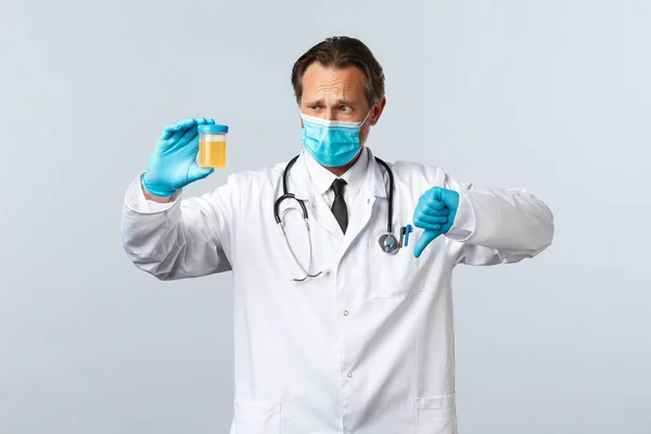 Covid-19, Prävention von Viren, Gesundheitspersonal und Impfkonzept. Enttäuschter Arzt in medizinischer Maske und Handschuhen zeigt Urinprobe, Daumen nach unten haben schlechtes Testergebnis, weißer Hintergrund — Stockfoto