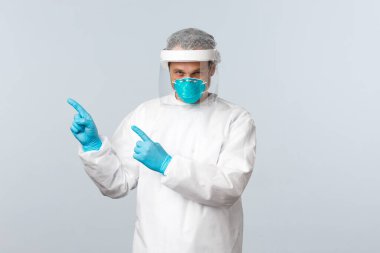 Covid-19, virüsü önleme, sağlık çalışanları ve aşı konsepti. Kişisel koruma ekipmanları içinde ciddi bir doktor. Sol üst köşeyi işaret ediyor. Bilgi panosuna.