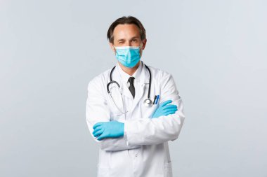 Covid-19, virüsü önleme, sağlık çalışanları ve aşı konsepti. Tıbbi maskeli ve eldivenli dost canlısı profesyonel doktor, hastayı rahatlatmaya özen göster, göz kırp ve kollarını çapraz bağla.