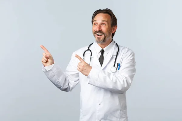 Covid-19, Coronavirus-Ausbruch, Gesundheitspersonal und Pandemiekonzept. Glücklich lächelnder Arzt in weißem Mantel zeigt amüsiert mit den Fingern in die linke obere Ecke und zeigt Medikamente oder Klinikbanner — Stockfoto