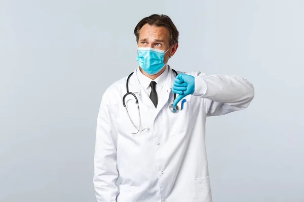 Covid-19: 바이러스 예방, 의료 종사자 및 예방 개념. 당황 한 의학박사가 마스크를 쓰고 장갑은 엄지 아래를 보여 주고 왼쪽 위 구석에 있는 프로 모를 보여 주고 있다. — 스톡 사진