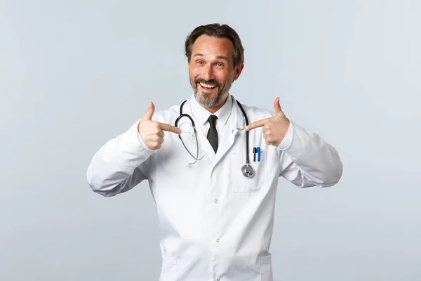 Covid-19, coronavirus outbreak, healthcare workers and pandemic concept. Happy professional male doctor in white coat promote his own clinic or services, pointing himself, bragging — Stock Photo, Image