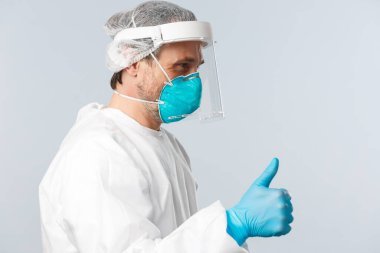 Covid-19, virüsü önleme, sağlık çalışanları ve aşı konsepti. Şahsi koruyucu ekipmanlı neşeli doktor profili baş parmak yukarı hastayı gösteriyor, her şeyin yolunda olduğundan emin olun, endişelenmeyin.