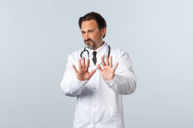 Covid-19, koronavirüs salgını, sağlık çalışanları ve pandemik konsept. İsteksiz ve hoşnutsuz beyaz önlüklü erkek doktor hayır diyor. Reddetme hareketi yap, durmasını ya da uzak durmasını söyle.