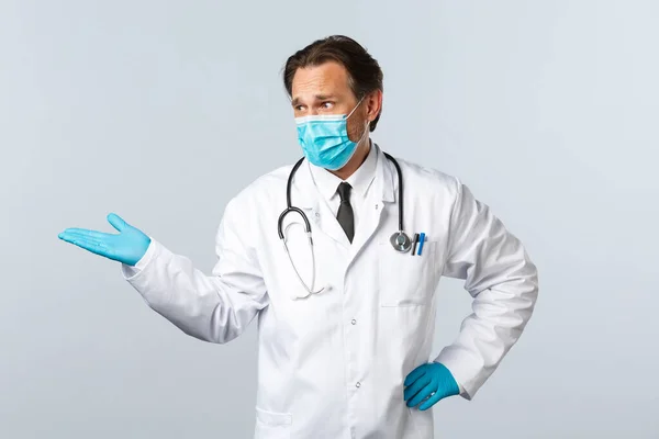 Covid-19, πρόληψη του ιού, εργαζόμενοι στον τομέα της υγείας και την έννοια του εμβολιασμού. Απογοητευμένος γιατρός με ιατρική μάσκα και γάντια, δείχνει και κοιτάζει κάτι κακό, λευκό φόντο. — Φωτογραφία Αρχείου