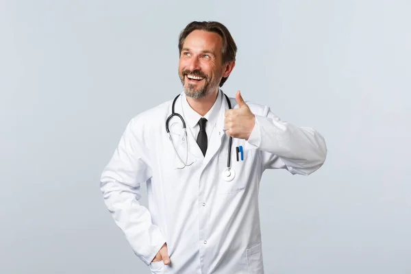 Covid-19, ξέσπασμα του ιού της στέψης, επαγγελματίες υγείας και πανδημία. Χαρούμενος όμορφος χαμογελαστός γιατρός με λευκό παλτό στην πάνω αριστερή γωνία, επιδείξτε την έγκρισή σας. — Φωτογραφία Αρχείου