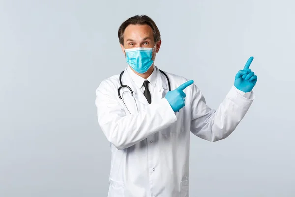 Covid-19, virüsü önleme, sağlık çalışanları ve aşı konsepti. Beyaz önlüklü, tıbbi maskeli ve eldivenli heyecanlı ve ilgi çekici erkek doktor sağ üst köşedeki klinik reklamını gösteriyor. — Stok fotoğraf