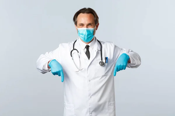 Covid-19, πρόληψη του ιού, εργαζόμενοι στον τομέα της υγείας και την έννοια του εμβολιασμού. Ευχάριστος γιατρός με ιατρική μάσκα και γάντια δίνουν συμβουλές, συστήνουν promo, δείχνοντας τα δάχτυλα κάτω στο πανό — Φωτογραφία Αρχείου