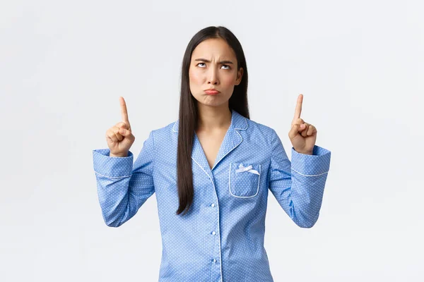 Complaining and bothered asian girl pouting and frowning displeased, looking and pointing fingers up disappointed, standing in blue pajamas, standing unsatisfied white background — Stock Photo, Image