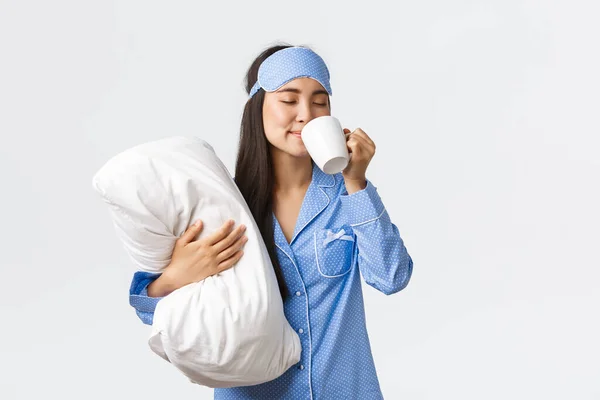 Morning lifestyle, breakfast and people concept. Smiling beautiful asian girl in pyjamas and sleeping mask, hugging pillow, having coffee in bed and watching tv, home leisure on weekends — Stock Photo, Image