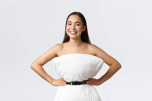 Beautiful asian girl making dress from pillow, secure it with belt around waste and posing for social media, smiling pleased, standing white background delighted