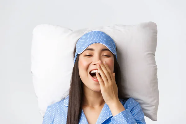 Крупный план счастливой улыбающейся азиатской девушки, лежащей в постели на подушке в голубой пижаме и в масках для сна, зевающей довольной после хорошего ночного сна, позирующей на белом фоне — стоковое фото