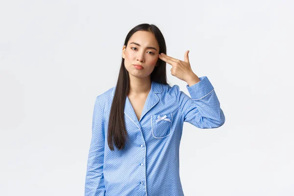 Irritada e incomodada menina asiática em pijama azul olhando com relutância, atirando-se com gesto arma como se sentindo farto, cansado de ouvir ou ver algo chato ou burro, fundo branco — Fotografia de Stock