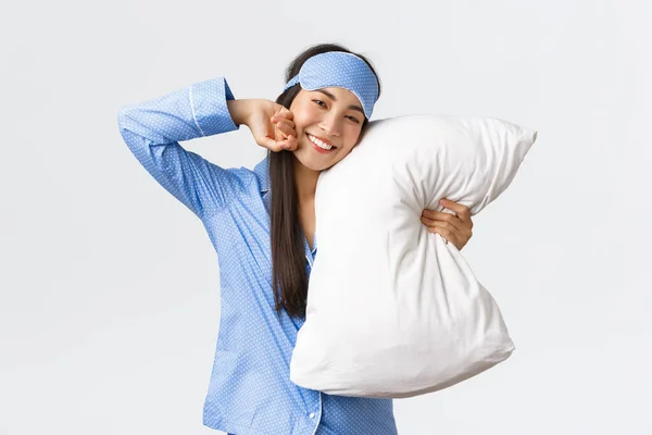 Zadowolona piękna azjatycka dziewczyna w niebieskiej piżamie i masce do spania, leżąca w łóżku i przytulająca poduszkę, uśmiechnięta z satysfakcji jak rozciąganie i dobre samopoczucie po nocnym śnie, białe tło — Zdjęcie stockowe