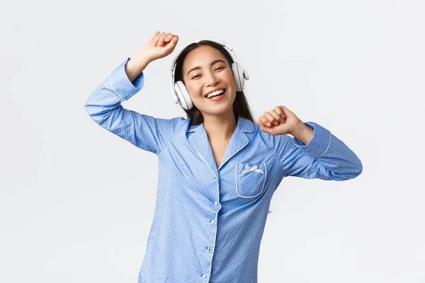 Casa lazer, fins de semana e conceito de estilo de vida. Alegre menina asiática feliz no pijama se divertindo, dançando à música em fones de ouvido, ouvindo música favorita no dia-de-folga, de pé alegre fundo branco — Fotografia de Stock