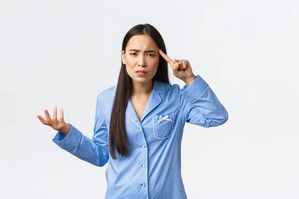 Molestado y confundido chica asiática quejándose de alguien actuando estúpido o loco, usando pijama azul, levantando la mano frustrado y rodar el dedo sobre el templo con consternación, regañando a alguien — Foto de Stock