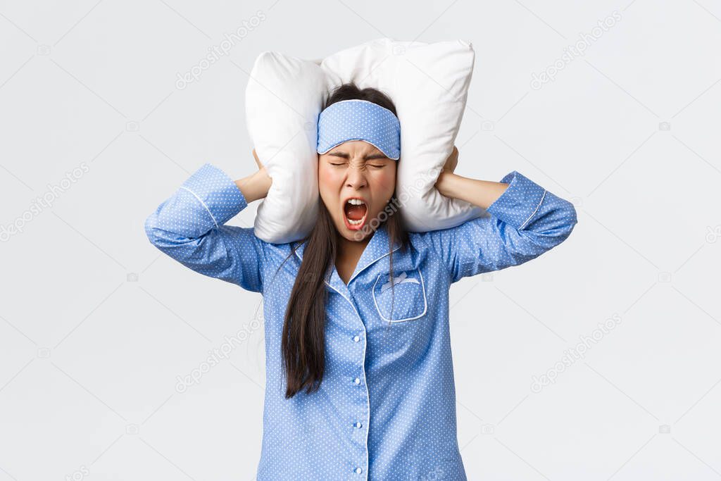 Angry furious asian girl in pajamas and sleeping mask, lying bed and shut ears with pillow, shouting mad as cant sleep from loud noise, neighbours through party at night, complaining annoying sound