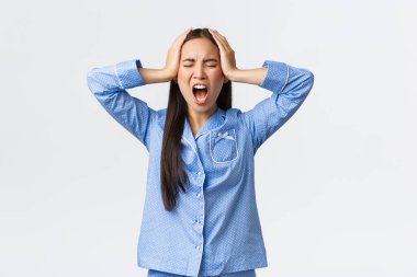 Distressed pissed-off asian girl in blue pajamas screaming outraged, holding hands on head in denial, got huge trouble, feeling upset and disappointed, standing white background