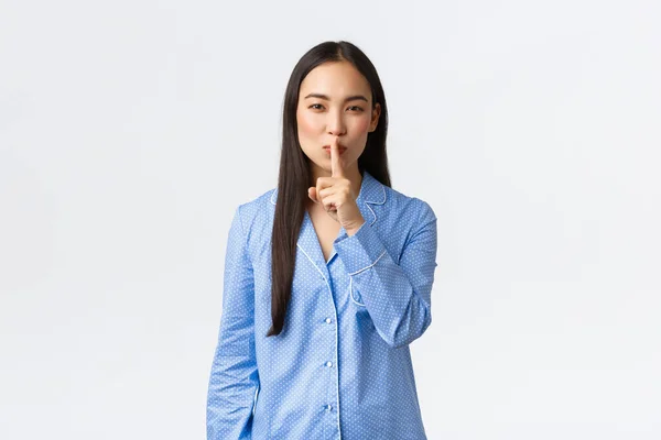 Sonriendo astuta linda chica asiática en pijama azul organizar sorpresa secreta para las novias de pijamada, mostrando gesto de silencio, silenciar quieren silencio, pidiendo silencio, de pie sobre fondo blanco —  Fotos de Stock