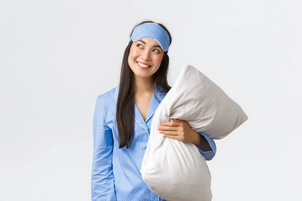 Przebiegła i troskliwa Kawaii azjatycka dziewczyna w niebieskiej piżamie i masce do spania, trzymająca poduszkę i wyglądająca ciekawie w lewym górnym rogu, uśmiechnięta przebiegła jak mająca pomysł, obrazująca coś, białe tło — Zdjęcie stockowe
