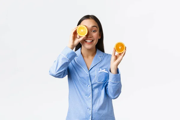 Matin, mode de vie actif et sain et concept de la maison. Souriant belle femme asiatique en pyjama bleu montrant des oranges et regardant optimiste, préparer du jus frais pour commencer la journée, fond blanc — Photo