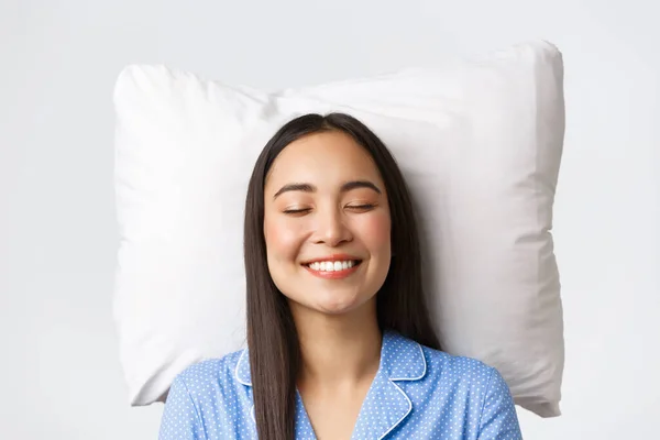 Sorridente sognante e bella donna asiatica sdraiata sul letto sul cuscino, indossa pigiami blu, chiude gli occhi e sorride, sogna ad occhi aperti o dorme di notte, imaging qualcosa di carino, sfondo bianco — Foto Stock