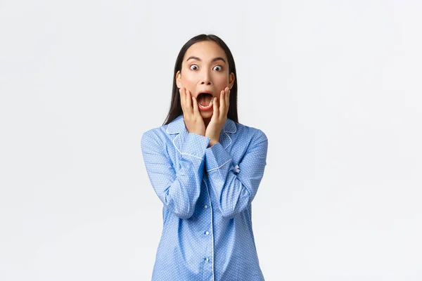 Překvapená a šokovaná asijská dívka v modrém pyžamu křičící v úžasu a úžasu, držící se za ruce nad otevřenými ústy a zírající na ohromenou kameru, stojící na bílém pozadí — Stock fotografie