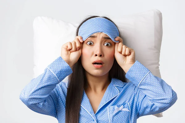 Потрясенная и испуганная беспокойная азиатка в синей пижаме, лежащая в постели на подушке, снимающая маску для сна и испуганная перед камерой, паникующая, как будто что-то забыла, белый фон — стоковое фото