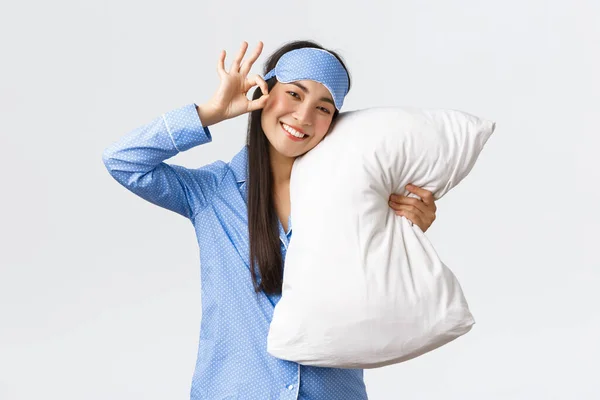 Nöjd glad leende och söt asiatisk flicka i blå pyjamas och sovande ögonmask, krama kudde i mask, visar okej gest, rekommendera produkt eller medicin för sömn, vit bakgrund — Stockfoto