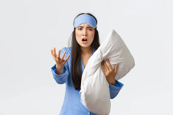 Angry bothered asian girl with insomnia, wearing sleeping mask and pajama, looking pissed-off as holding pillow and shaking hand furious, complaining on noise at night, white background — Stock Photo, Image