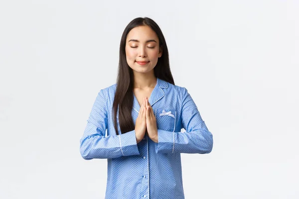 Leende avslappnad och lugn asiatisk flicka i blå pyjamas blunda och hålla händerna tillsammans i vädjan, att önska, be innan du går och lägger dig, drömmer och står över vit bakgrund — Stockfoto