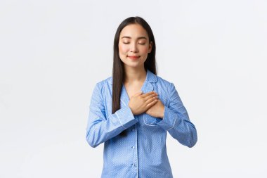 Rüya gibi ve dokunaklı mavi pijamalı Asyalı kadın, kapalı gözler ve mutlu bir gülümsemeyle kalbe dokun, bir şeyi hatırla, tatlı anılarını içinde tut, beyaz arka plan üzerinde pijamalar içinde duran