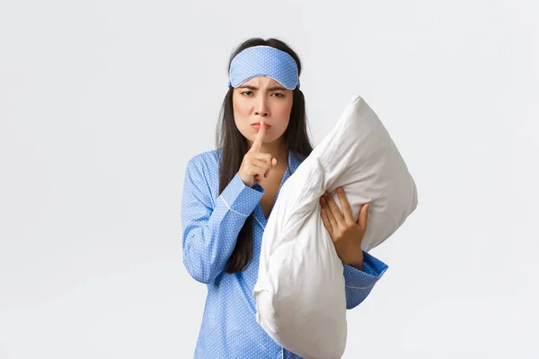Angry displeased asian girl in sleeping mask and pyjamas, holding pillow and frownign disturbed, shushing, say shhh as woke-up from loud noise, asking people keep quiet at night, white background — Stock Photo, Image