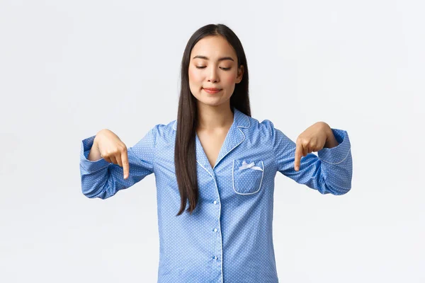 Agradável menina asiática em pijama azul vendo algo tentador, sorrindo satisfeito e pensativo, apontando os dedos para baixo no banner inferior com o produto, encontrou algo bom, fundo branco — Fotografia de Stock