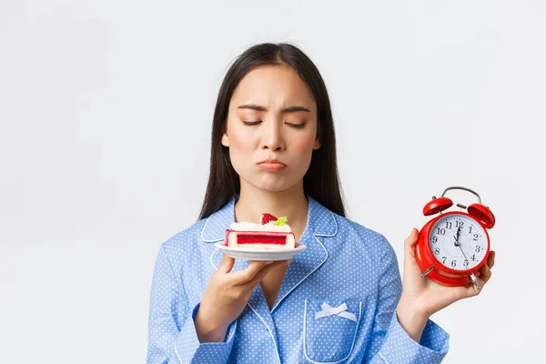 Close-up of upset cute pouting asian girl in pajama showing clock and looking with regret and temptation at tasty piece of cake, wanting dessert but its too late, standing white background