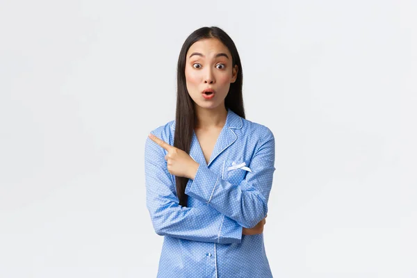 stock image Surprised pretty young asian girl look amazed at camera while pointing finger left at awesome new product. Woman in blue pajamas showing something cool, standing white background