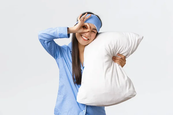 Glädjande entusiastisk vacker asiatisk flicka i blå pyjamas och sovmask, krama kudde och leende upbeat som går sängen, redo för övernattning, visar okej gest i godkännande, vit bakgrund — Stockfoto