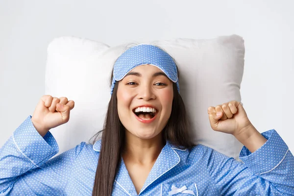 Close-up of enthusiastic asian girl in blue pajamas and sleeping mask, stretching hands up delighted after good night sleep, take-off eyemask in morning, lying in bed on pillow and smiling happy — Stock Photo, Image