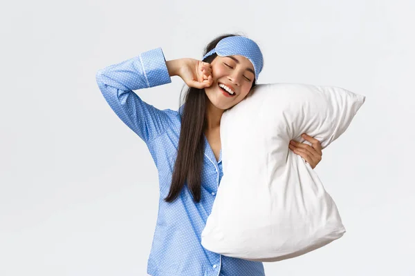 Leende glad söt asiatisk flicka i blå pyjamas och sovmask, krama kudde och stretching händer glad som äntligen gå sängen, vill sova eller vakna på morgonen, vit bakgrund — Stockfoto