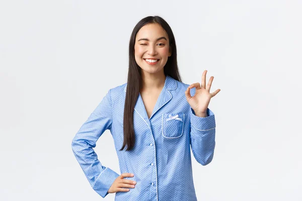 Mavi pijamalı mutlu Asyalı kız göz kırpıyor ve mutlu bir gülümsemeyle iyi kalite tavsiye ediyor, mükemmel hizmeti garanti ediyor, beyaz arka planda tatmin oluyor. — Stok fotoğraf