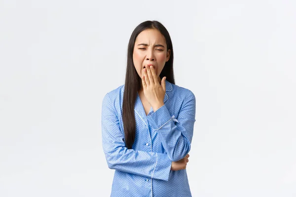 Fáradt és unatkozó aranyos ázsiai lány kék pizsamában ásítozik unalmas történetekből az ottalvós bulin, kimerült, érdeklődés hiányát mutatja, fehér háttér fölött áll aludni akar menni — Stock Fotó